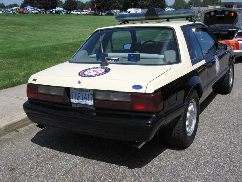 1991 Ford LTD Crown Victoria - Rear three-quarter view of a black-and-tan Ford Mustang LX police coupe with a circular patrol badge decal at a car show.