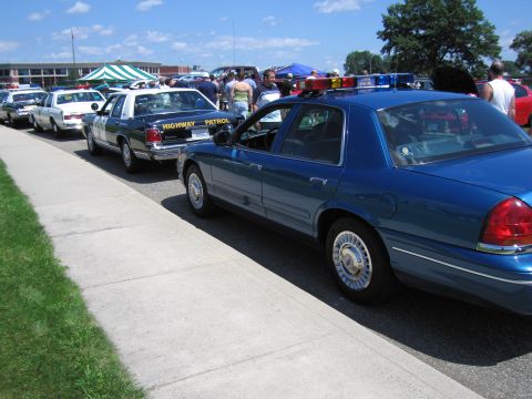 1991 Ford LTD Crown Victoria - row of parked police cars including a blue Ford Crown Victoria and a Highway Patrol sedan with crowds at an outdoor show.
