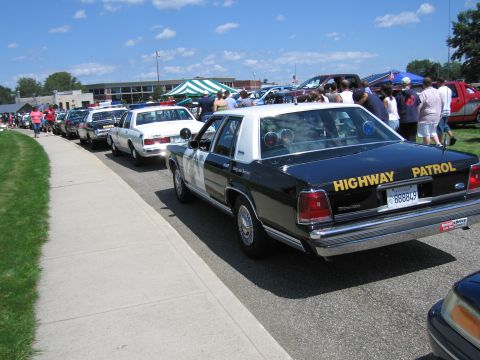 1991 Ford LTD Crown Victoria - Rear view of a black-and-white Highway Patrol Ford sedan leading a long row of police cars past crowds at an outdoor show.