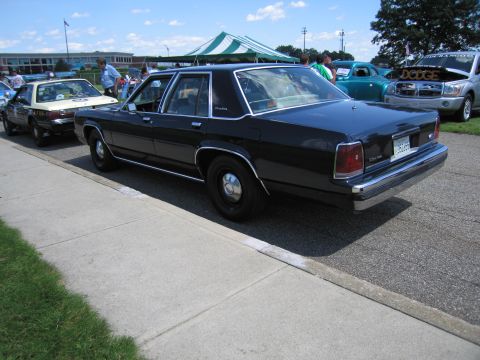 1991 Ford LTD Crown Victoria - Rear three-quarter view of a dark blue Ford sedan with steel wheels parked in a row of cars at an outdoor show.