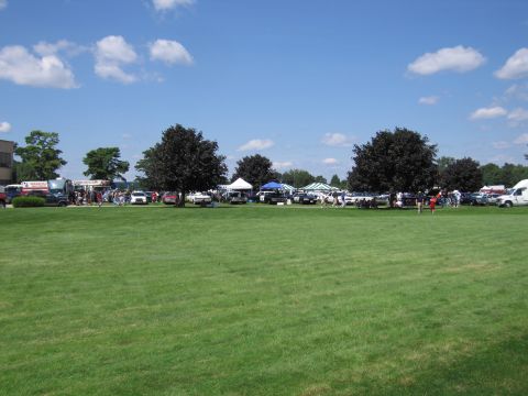 1991 Ford LTD Crown Victoria - wide green field with distant tents, parked cars, and crowds under a blue sky with scattered clouds at an outdoor event.