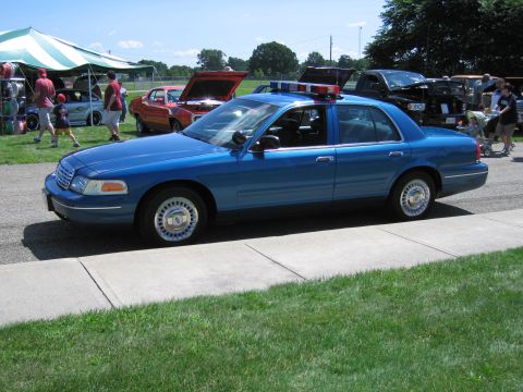 1991 Ford LTD Crown Victoria - Side view of a blue Ford Crown Victoria police sedan with a roof light bar parked near tents at an outdoor car show.