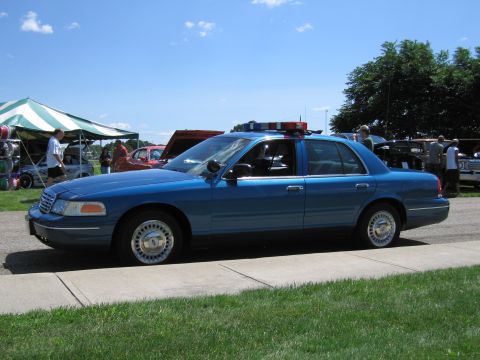 1991 Ford LTD Crown Victoria - Driver-side profile of a blue Ford Crown Victoria police sedan with a roof light bar parked on pavement near show tents.