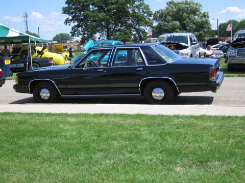 1991 Ford LTD Crown Victoria - plain black full-size Ford sedan parked on pavement beside a grassy field at an outdoor car show.