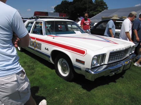 1991 Ford LTD Crown Victoria - white Plymouth State Police station wagon with red and blue striping and roof light bar among onlookers at a car show.