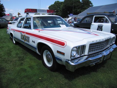 1991 Ford LTD Crown Victoria - white Plymouth police sedan with red and blue body stripes and roof light bar displayed on grass at a car show.
