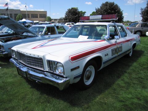 1991 Ford LTD Crown Victoria - white Plymouth police sedan with red and blue striping, whitewall tires and roof light bar parked on grass at a car show.