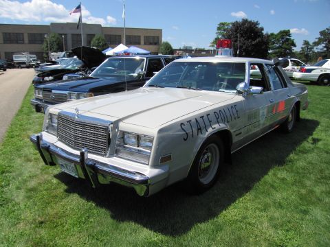 1991 Ford LTD Crown Victoria - silver Chrysler State Police sedan with red stripe, push bar and roof light parked on grass at an outdoor car show.