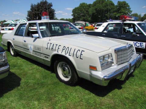 1991 Ford LTD Crown Victoria - cream-colored Rhode Island State Police sedan with chrome push bar and roof light bar displayed on grass at a car show.