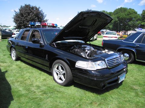 1991 Ford LTD Crown Victoria - black Ford Crown Victoria police interceptor with raised hood and roof light bar parked on grass at a car show.