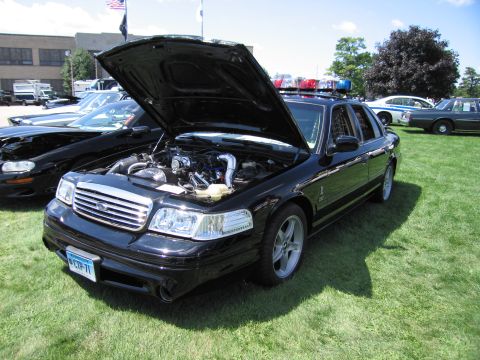 1991 Ford LTD Crown Victoria - black Ford police sedan with hood raised exposing the engine and a roof light bar, displayed on grass at a car show.
