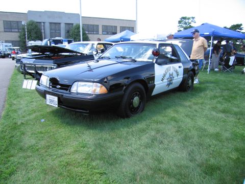 1991 Ford LTD Crown Victoria - black-and-white Highway Patrol Ford Mustang 5.0 coupe with roof light parked on grass at an outdoor car show.