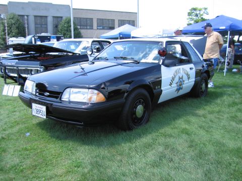 1991 Ford LTD Crown Victoria - black-and-white Highway Patrol Ford Mustang 5.0 with star door shield and roof light displayed on grass at a car show.