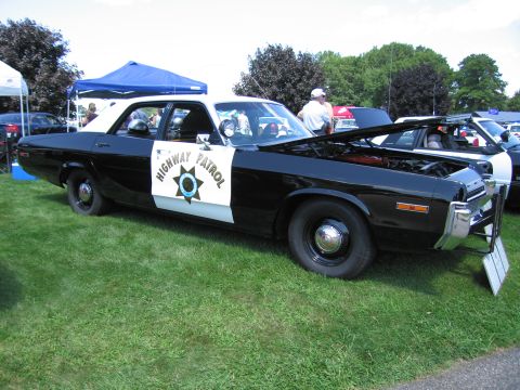 1991 Ford LTD Crown Victoria - black-and-white Highway Patrol Dodge sedan with star door shield and raised hood displayed on grass at a car show.