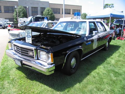 1991 Ford LTD Crown Victoria - black-and-white Ford LTD police sedan with door shield emblem, number 45 and raised hood parked on grass at a show.