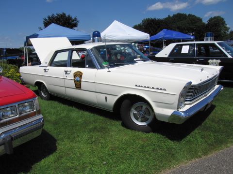 1991 Ford LTD Crown Victoria - white Ford Galaxie 500 police sedan with door shield, roof light and raised hood parked on grass at a car show.