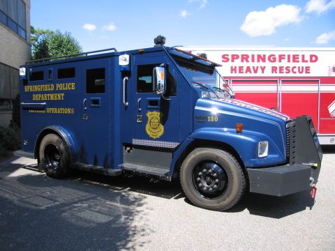 1991 Ford LTD Crown Victoria - blue Springfield Police Department armored tactical truck, unit 130, parked next to a red Springfield Heavy Rescue apparatus.
