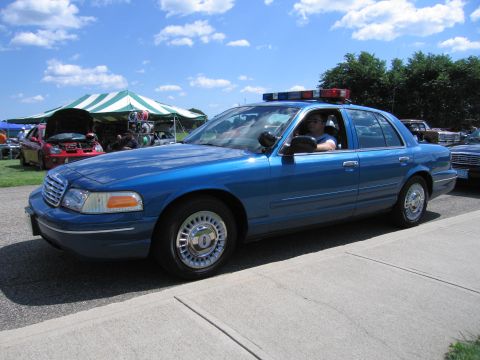 1991 Ford LTD Crown Victoria - blue Ford Crown Victoria police sedan with a roof light bar parked on pavement at a car show under blue sky.