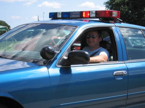 1991 Ford LTD Crown Victoria - man wearing sunglasses sits at the wheel of a blue State Police car with an illuminated roof light bar.