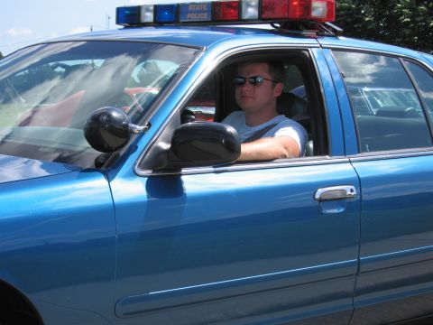 1991 Ford LTD Crown Victoria - man in sunglasses drives a blue State Police sedan, his arm resting on the door, light bar lit on the roof.