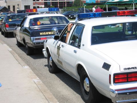1991 Ford LTD Crown Victoria - lineup of vintage police cars along a curb, led by a white Chevrolet with a black Highway Patrol sedan behind.