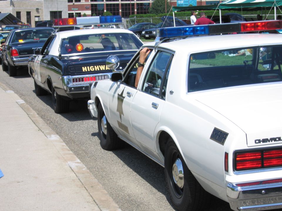 Row of classic police cars including a white Chevrolet and a black Highway Patrol sedan