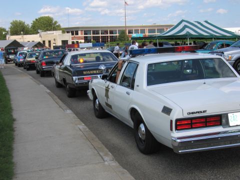 1991 Ford LTD Crown Victoria - row of vintage police cars parked along a sidewalk at a show, with tents and a building in the background.