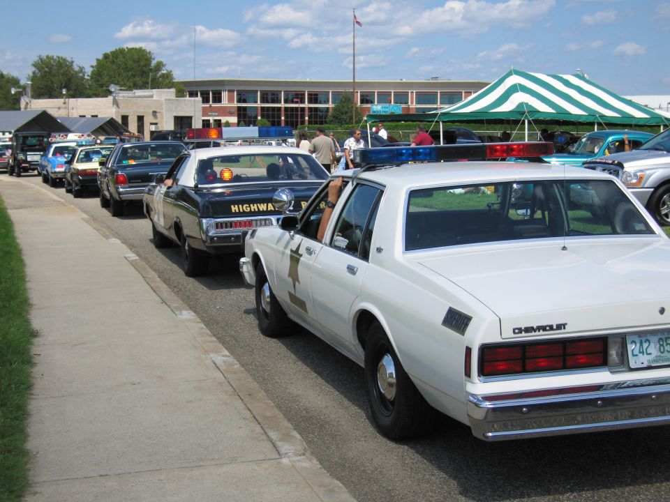 White Chevrolet police car and black Highway Patrol sedan lined up at a car show