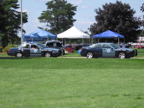 1991 Ford LTD Crown Victoria - State Police sedan and a Dodge Charger police cruiser parked on a lawn beside event tents and trees.