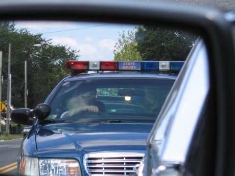 1991 Ford LTD Crown Victoria - blue State Police Ford with an illuminated light bar follows on a tree-lined road, seen through a rear window.