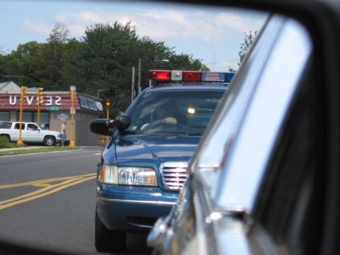 1991 Ford LTD Crown Victoria - blue police car with a roof light bar waits at an intersection near a 7-Eleven, viewed past another vehicle.