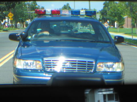 1991 Ford LTD Crown Victoria - blue Ford police sedan with a lit roof light bar on a tree-lined road, viewed through a windshield.