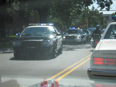 1991 Ford LTD Crown Victoria - Several police cars with lit lights and a motorcycle officer move along a street, viewed through a windshield.