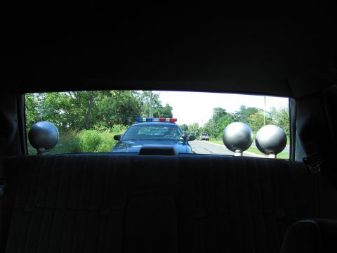 1991 Ford LTD Crown Victoria - Looking out the rear window of a car past spotlights at a blue police car with light bar following on the road.