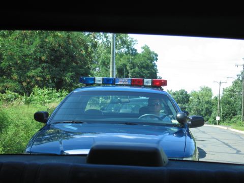 1991 Ford LTD Crown Victoria - blue State Police Ford with a roof light bar travels a country road, seen through a car's rear window.