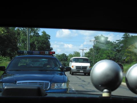 1991 Ford LTD Crown Victoria - blue police car with light bar and a white van on a road, viewed through a rear window past chrome spotlights.