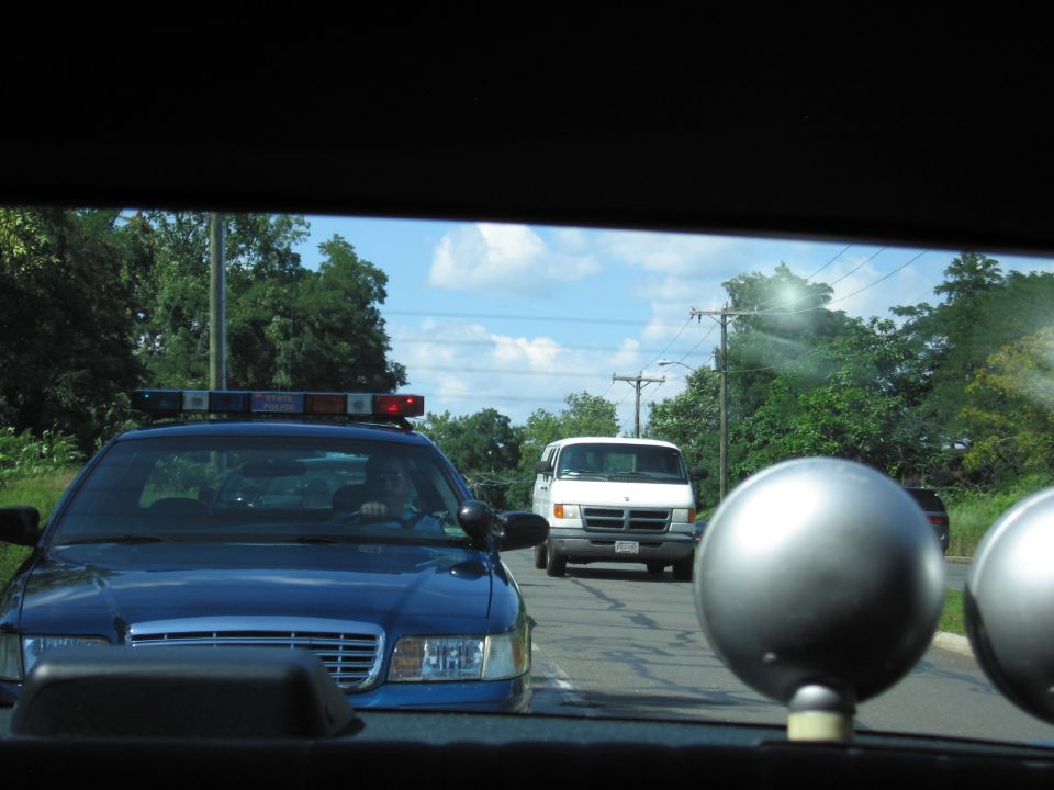 Blue police car and a white van on a road seen through a rear window with spotlights