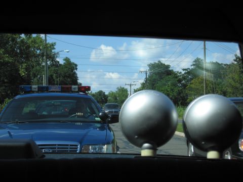 1991 Ford LTD Crown Victoria - blue Ford police car with a roof light bar follows on a road, framed by two chrome spotlights through a rear window.