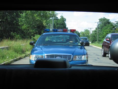 1991 Ford LTD Crown Victoria - blue Ford Crown Victoria police car with a roof light bar travels a road, seen through the rear window of a car.