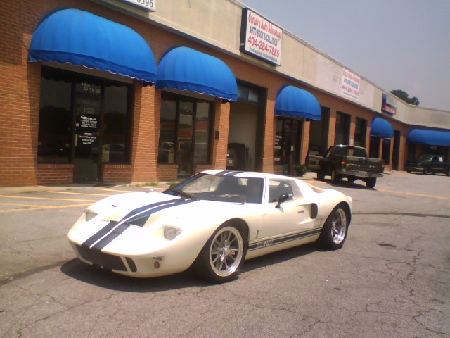 White Ford GT-style supercar with blue stripes parked outside brick auto body shop bays