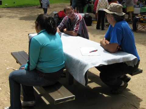 2003 Ford Taurus - Three people sit talking around a picnic table covered with a white cloth at an outdoor gathering, with a clipboard and pen on the table an…