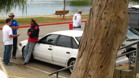 2003 Ford Taurus - Group of men talking next to a white sedan in a parking lot near a lake and tree.