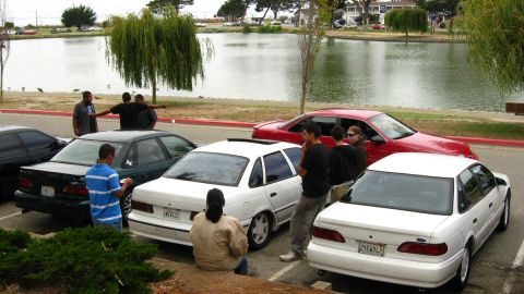 2003 Ford Taurus - A group of people stand near several sedans parked in a lot next to a body of water.