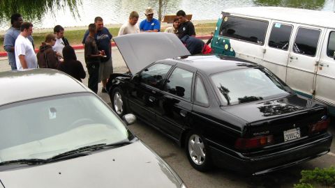 2003 Ford Taurus - A black second-generation Ford Taurus with its hood up and California plate 2XXU425 sits at a lakeside meet on white five-spoke wheels, vie…