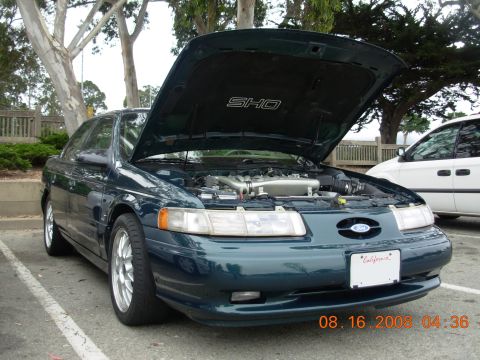 1995 Ford Taurus - A dark green sedan, possibly a Ford Taurus SHO, is parked with its hood open, revealing the engine and an "SHO" emblem on the underside of…