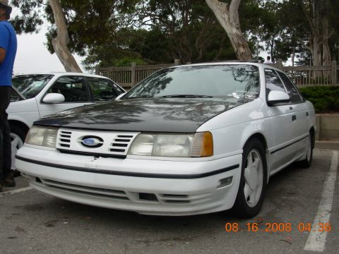 1995 Ford Taurus - A white Ford Taurus with a black hood is parked in a lot, with another car visible to its left.