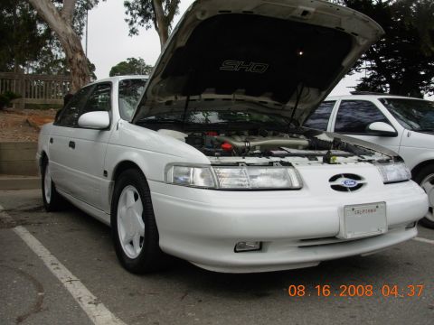 1995 Ford Taurus - A white 1995 Ford Taurus SHO is parked with its hood open, revealing the engine bay. Another Taurus is visible to the right.