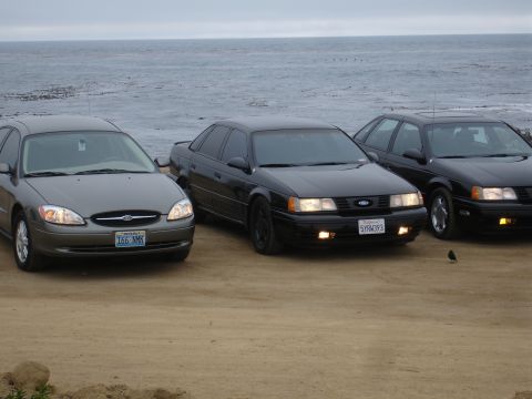1990 Ford Taurus - Three Ford Tauruses are parked near the ocean, with two black models and one grey model visible.