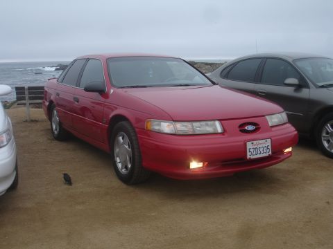 1990 Ford Taurus - A red sedan is parked near the ocean with another vehicle beside it under an overcast sky.
