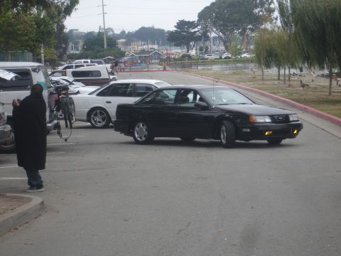 1990 Ford Taurus - A black sedan drives past other vehicles in a parking lot near a marina.
